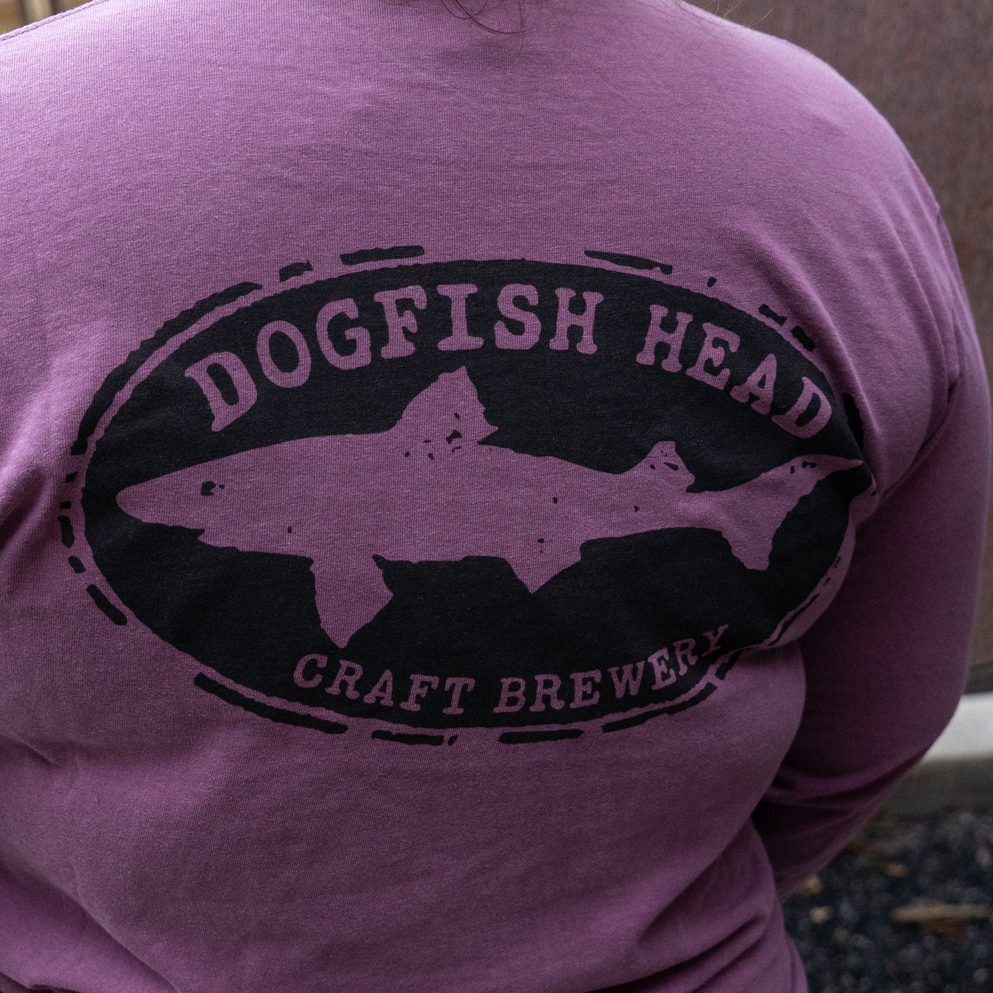 A person wearing a Berry Oversize Logo Long Sleeve from GO WEST T-SHIRT COMPANY, featuring a relaxed fit and a black Dogfish Head Craft Brewery shark logo on the back.