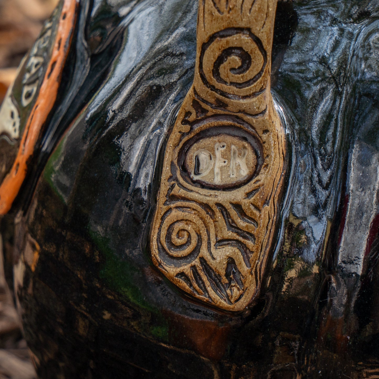A close-up of the Black Punkin Growler by Romanick Pottery: a glossy, dark ceramic vessel with a tan textured handle, intricate swirl carvings, and “DEN” engraved at the center—one of our limited edition growlers.