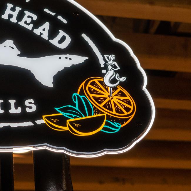The Merchtank Canned Cocktails LED Sign features a neon orange slice with green leaves, a mermaid, partial white text, a fish silhouette, and subtle Dogfish Head branding on a dark background.