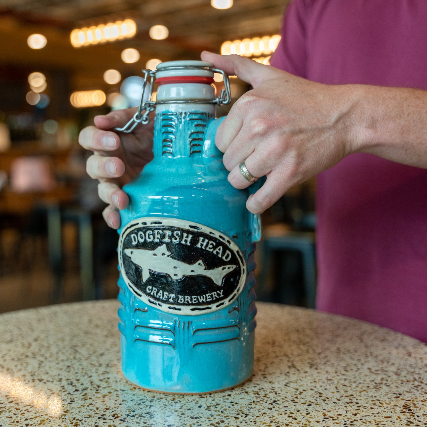 A person in a maroon shirt holds a large Romanick Pottery Coastal Blue Ceramic Growler with a wide-mouth hinge top, standing at a speckled tabletop in a well-lit indoor space.
