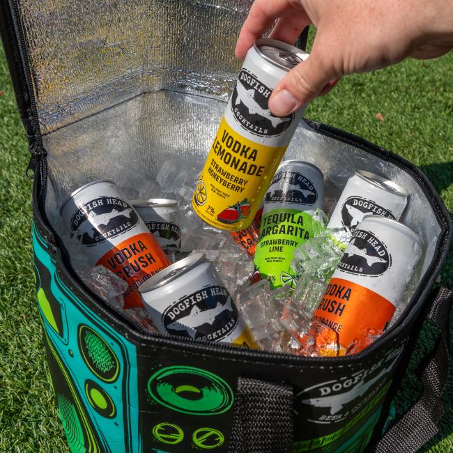A hand lifts a canned cocktail from a Go Match Up Cooler Bag filled with ice and drinks. The insulated soft cooler rests on green grass outdoors, keeping beverages perfectly chilled.