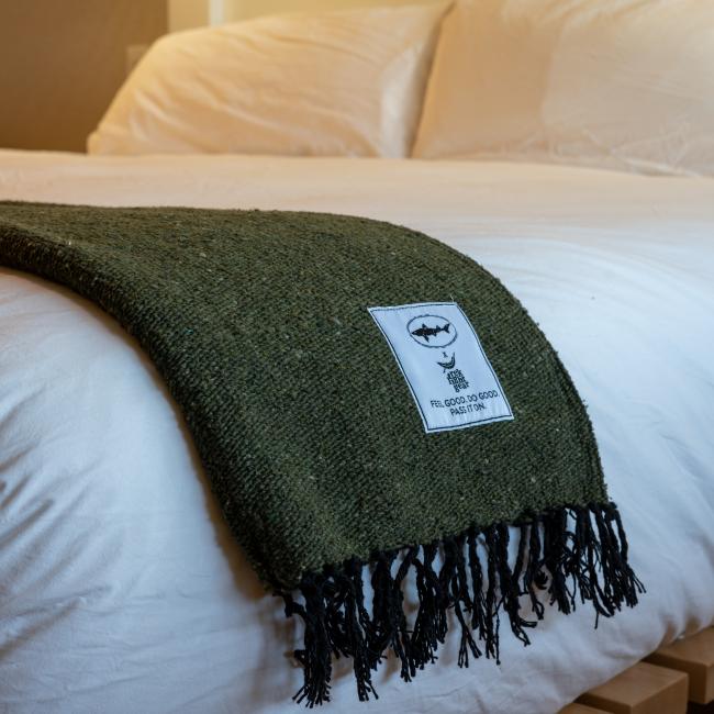 A green INN Blanket by Trek Light Gear with black fringe is draped over a neatly made bed with white bedding. The blanket has a white label featuring a bat illustration and text.