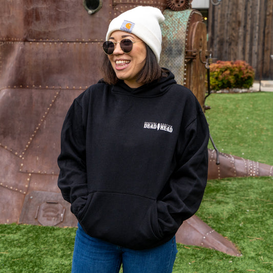 A smiling person wearing the Dogfish Head x Grateful Dead Black Hoodie by GO WEST T-SHIRT COMPANY, along with a white beanie, sunglasses, and blue jeans, stands on grass outdoors near a large metallic structure and wooden fence.