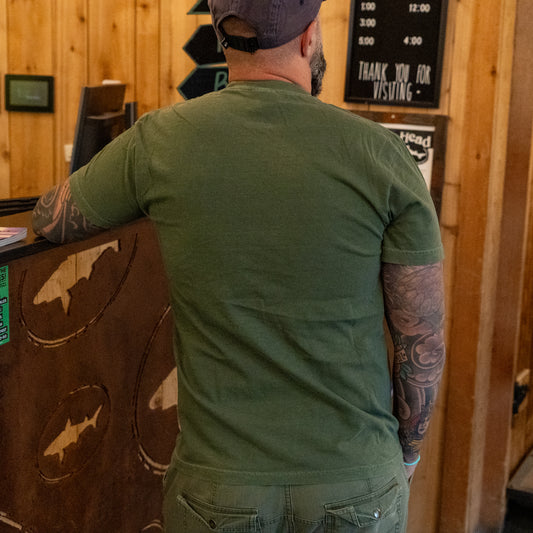 Wearing a Dogfish Head x Grateful Dead Green Logo Tee by GO WEST T-SHIRT COMPANY, a tattooed individual stands with their back to the camera at a rustic wooden counter, also sporting a green cap.