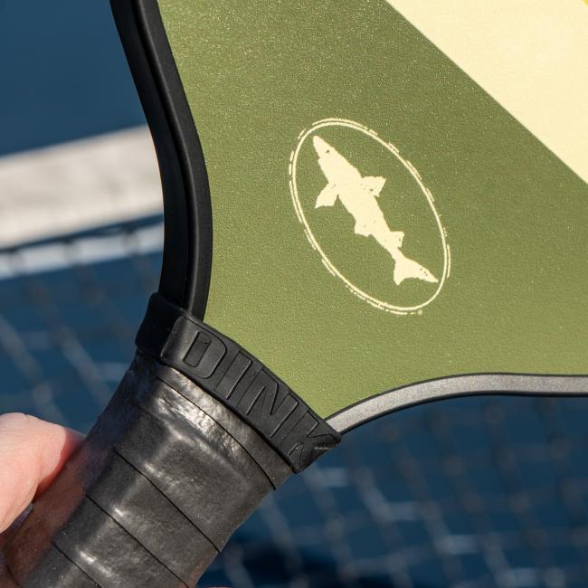 A close-up of the Dogfish Head Pickleball Paddle by Custom Pickleball - MAH Tech shows its black grip, olive green face, white shark logo, and honeycomb core, held in hand with a blurred net and court in the background.