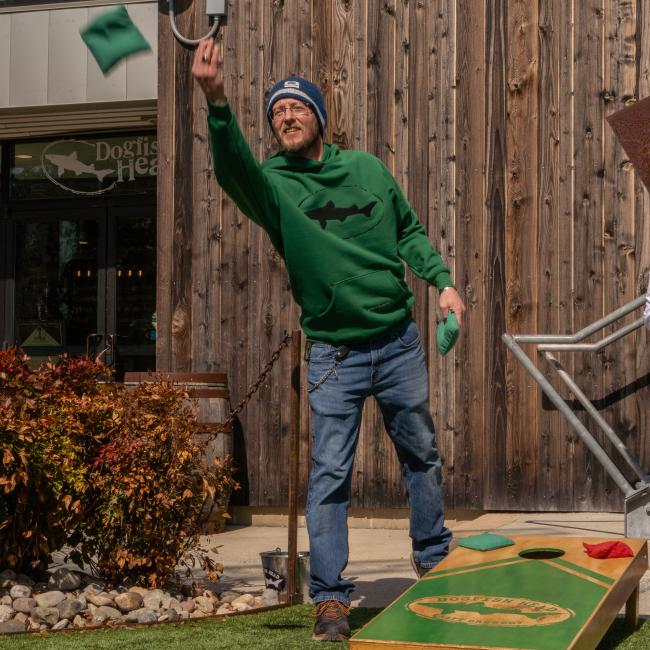 Wearing a Dogfish Head x Boathouse Green Heavyweight Hoodie by Boathouse Apparel and jeans, a man tosses a bean bag playing cornhole outside on artificial grass near a wooden building.