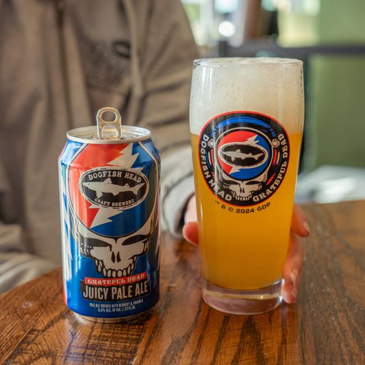 A person holds a Dogfish Head x Grateful Dead Glass by Grandstand Glassware & Apparel filled with hazy pale ale next to an open can on a wooden table. Both feature vibrant Grateful Dead-themed artwork.