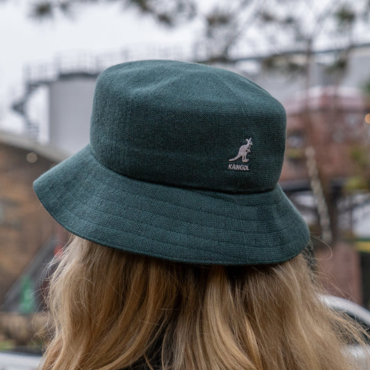 A person with long blonde hair is seen from behind wearing a dark green Dogfish Head x Grateful Dead KANGOL Bucket Hat by BOLLMAN HATS outdoors, with blurred buildings and trees in the background.