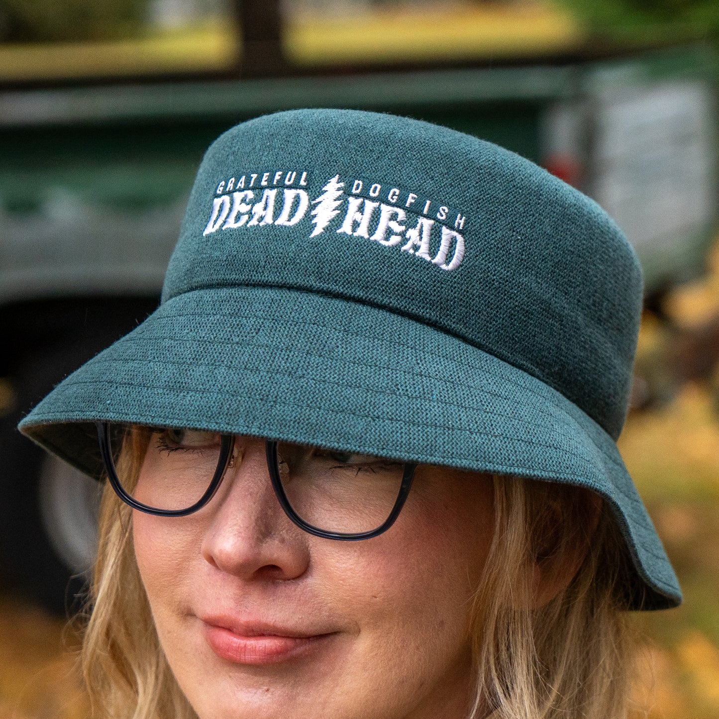 A person wearing glasses and a green Dogfish Head x Grateful Dead KANGOL Bucket Hat by BOLLMAN HATS with white embroidered text stands outdoors, with a blurred background featuring a vehicle and greenery.
