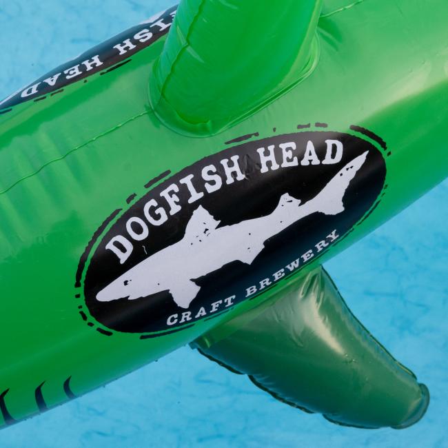 The Merchtank Dogfish Pool Float features a white dogfish shark silhouette on a black oval, with the Dogfish Head Craft Brewery logo, all on a green vinyl float perfect for pool fun.