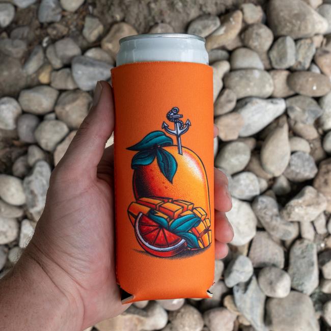 A hand holds a 12oz can in a slim orange Merchtank Four Slim Can Cooler featuring orange fruit, slices, and a silver anchor. Gray and brown rocks of various sizes are in the background.