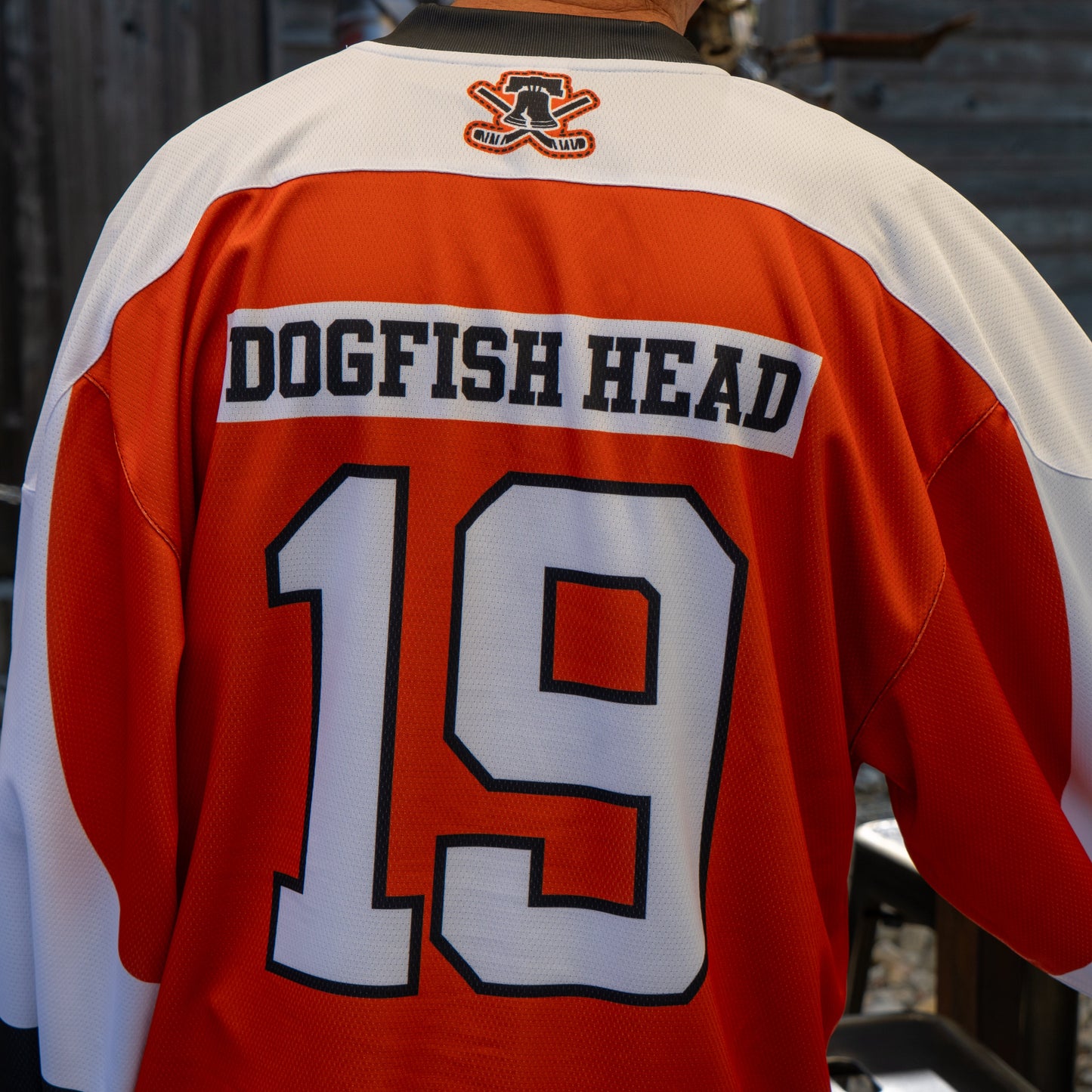 A person wears the Dogfish Head x Garnet Hathaway Engine 19 Hockey Jersey by In Your Face Apparel, featuring orange, white, and black colors with DOGFISH HEAD and 19 on the back; a logo is near the collar against a blurred background.