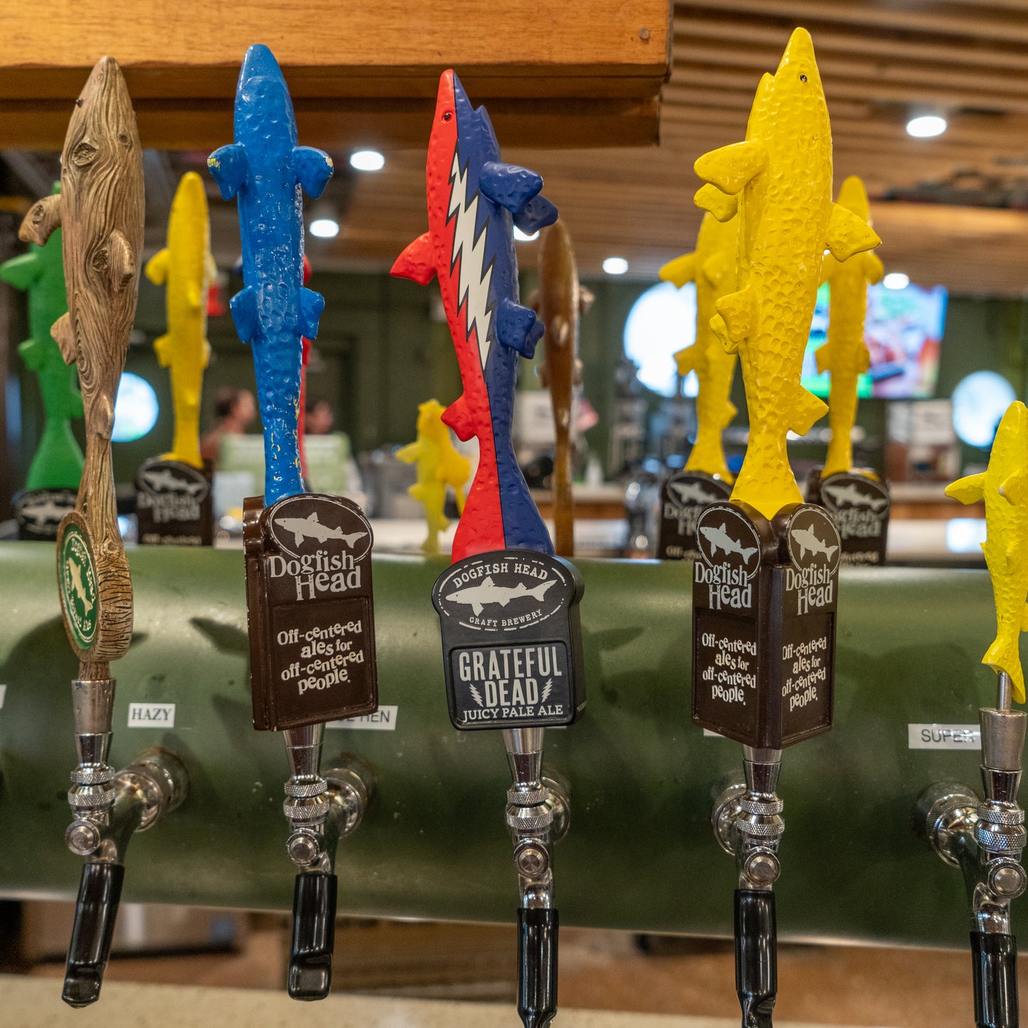 Close-up of colorful fish-shaped beer tap handles at a bar, including the Merchtank Dogfish Head x Grateful Dead Tap Handle with a red, white, and blue design featuring prominent logos and labels.