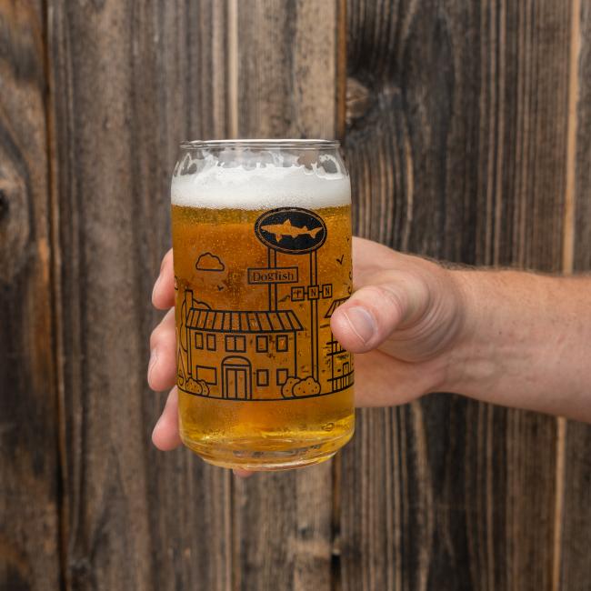 A hand holds a clear INN Can Glass by Grandstand Glassware & Apparel, filled with light beer and a foamy head. The glass displays an illustrated building, celebrating the Dogfish Head INN in Lewes, Delaware, against rustic wooden boards.