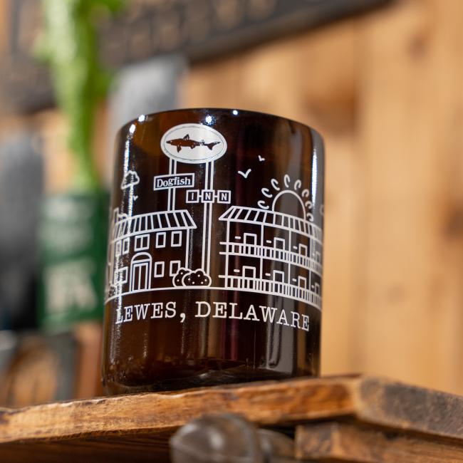The INN Growler Candle by Unwined Candles, LLC features a brown glass growler with white building and Dogfish Inn graphics, LEWES, DELAWARE text, double wicks, and a citrus scent. A slightly blurred wooden background is visible.