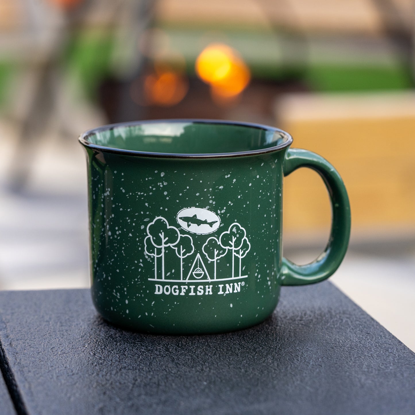 A green speckled 12oz Inn Camp Cup by Grandstand Glassware & Apparel, featuring Dogfish Inn with tree and fish graphics in white, sits on a dark outdoor surface amid blurred background lights.