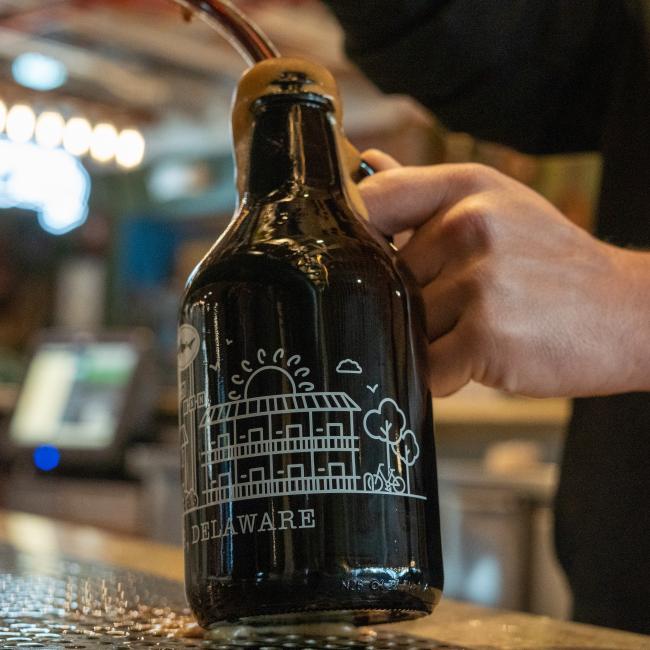 A person fills a Grandstand Glassware & Apparel Inn Growler 32oz, decorated with a white illustration of a building and trees reminiscent of the Dogfish Head INN, with beer from a tap at a bar or brewery.