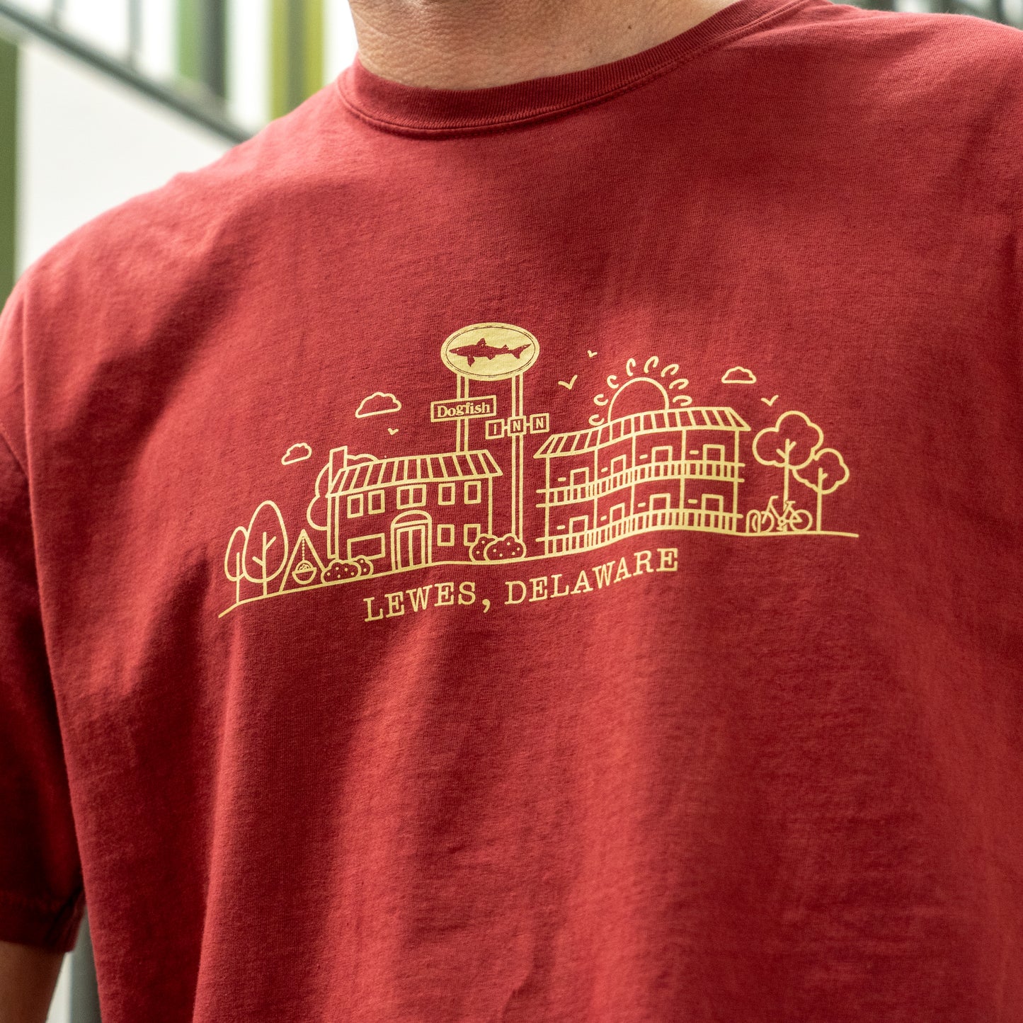 Wearing the Inn Red Line Art Tee by GO WEST T-SHIRT COMPANY, this red shirt features line art of buildings, trees, a sun, and a fish sign with “LEWES, DELAWARE,” capturing the craft spirit of this coastal town.