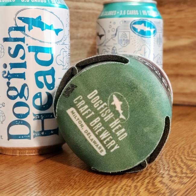 Three 12oz Dogfish Head cans sit on a wooden surface, one displaying the Dogfish Head Craft Brewery logo and "Milton, Delaware." Keep them chilled in your Off Centered Ales Treehouse Can Cooler from Grandstand Glassware & Apparel.