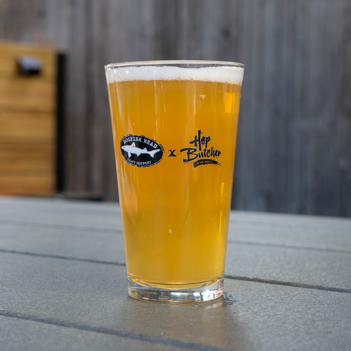 A pint glass of hazy golden beer rests on a gray outdoor table beside a can cooler, both part of the Crispy Slice Merch Bundle from GO WEST T-SHIRT COMPANY, with a blurred background of warm wood paneling.