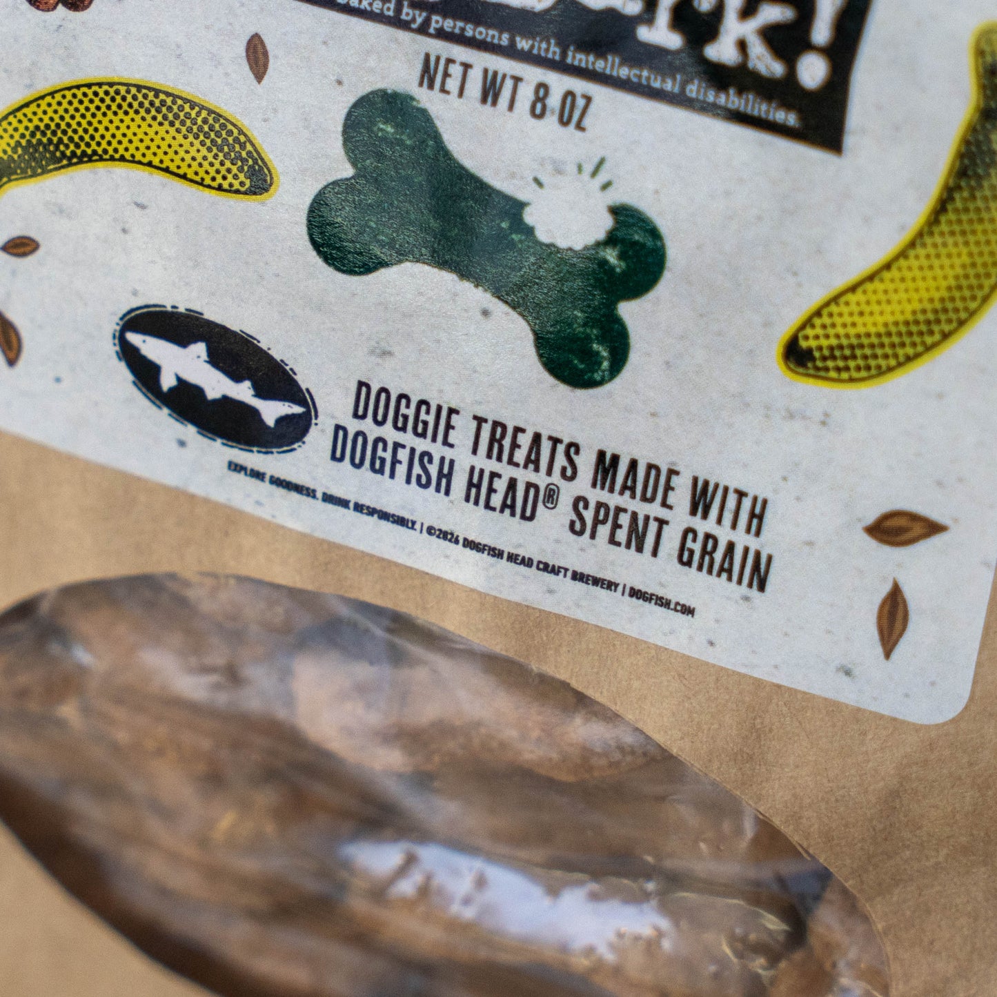 Close-up of a dog treat pouch label reading 'Doggie Treats Made with Dogfish Head Spent Grain' with banana and bone graphics, showing a clear window over brown treats inside.