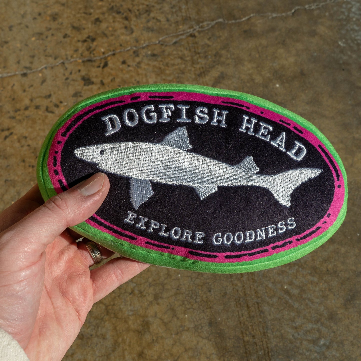 Hand holding an oval shaped Dogfish Head plush dog toy with a black logo that reads "Dogfish Head, Explore Goodness". The logo is outlined in hot pink and then outlined and covered in green.
