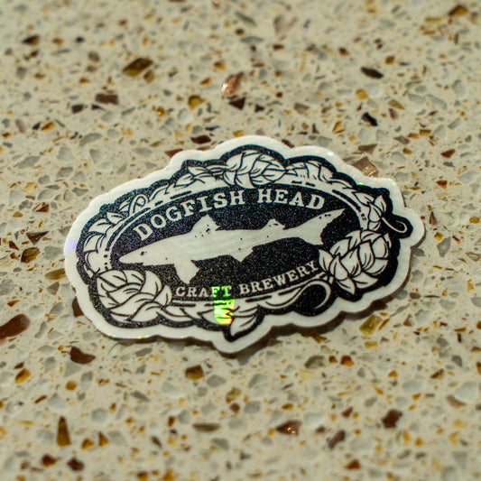 A black and white Rainbow Symphony Suncatcher Logo Sticker with a shark logo and leaf border, perfect for brewery gear fans, sits on a speckled beige countertop.