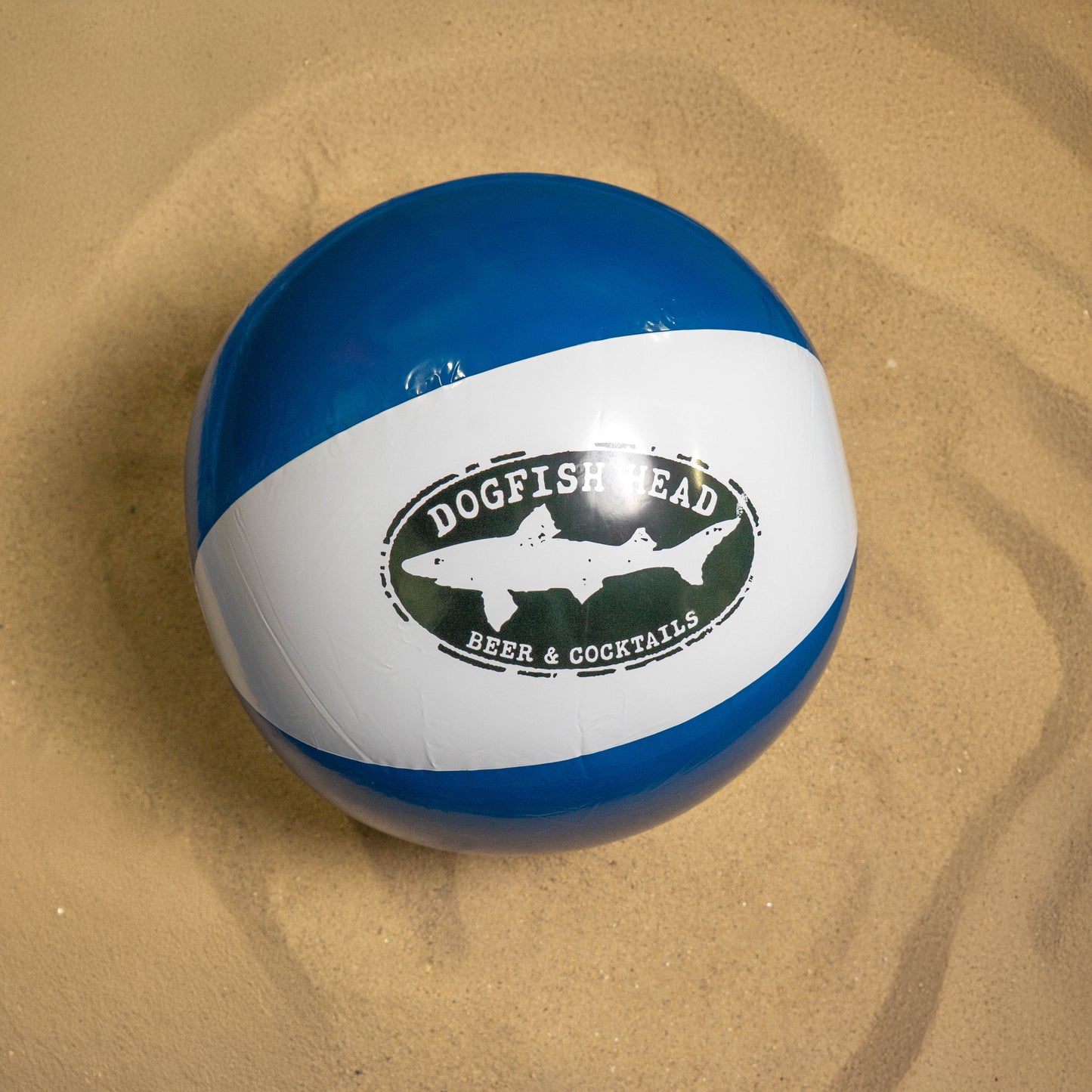 A blue and white beach ball with a Dogfish Head Beer & Cocktails logo, included in the Drink Summer Louder Festival Kit by Merchtank, rests on light sand—a fun summer festival essential.