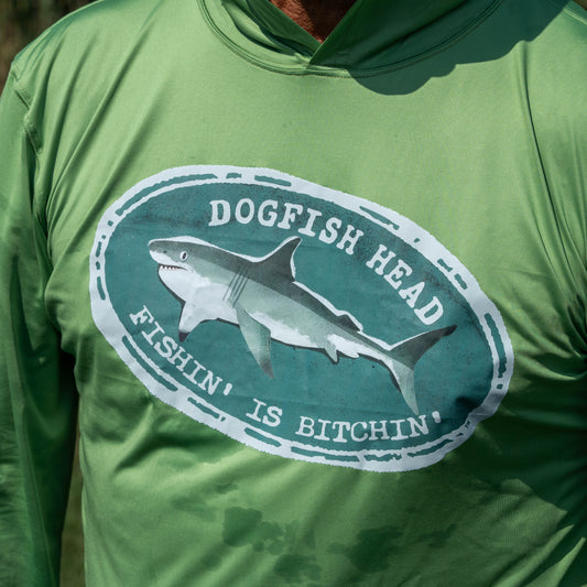 A person wearing the Boathouse Apparel Dogfish Head x Boathouse Fishin' is Bitchin' Sun Pullover Hoodie, a green moisture-wicking shirt featuring an oval logo with a shark and the words "Dogfish Head Fishin' Is Bitchin'" from the collaborative collection.