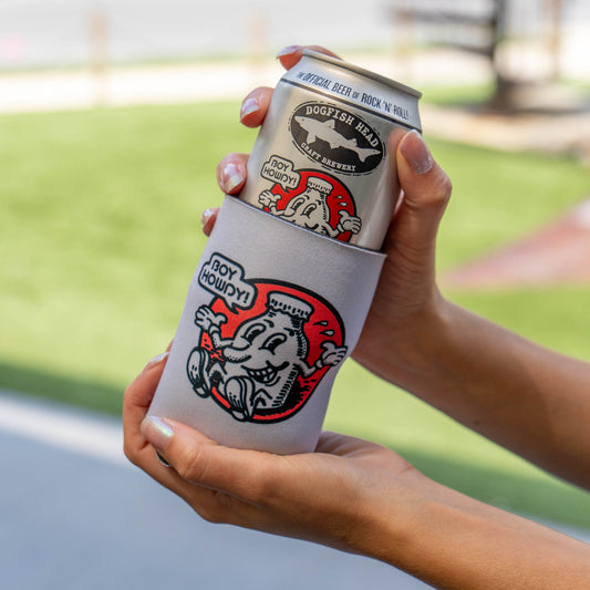 A person holds a silver can of Dogfish Head Beer in a light gray can cooler sleeve with a red and black cartoon graphic. The background is a blurred outdoor setting with green grass and a paved path.