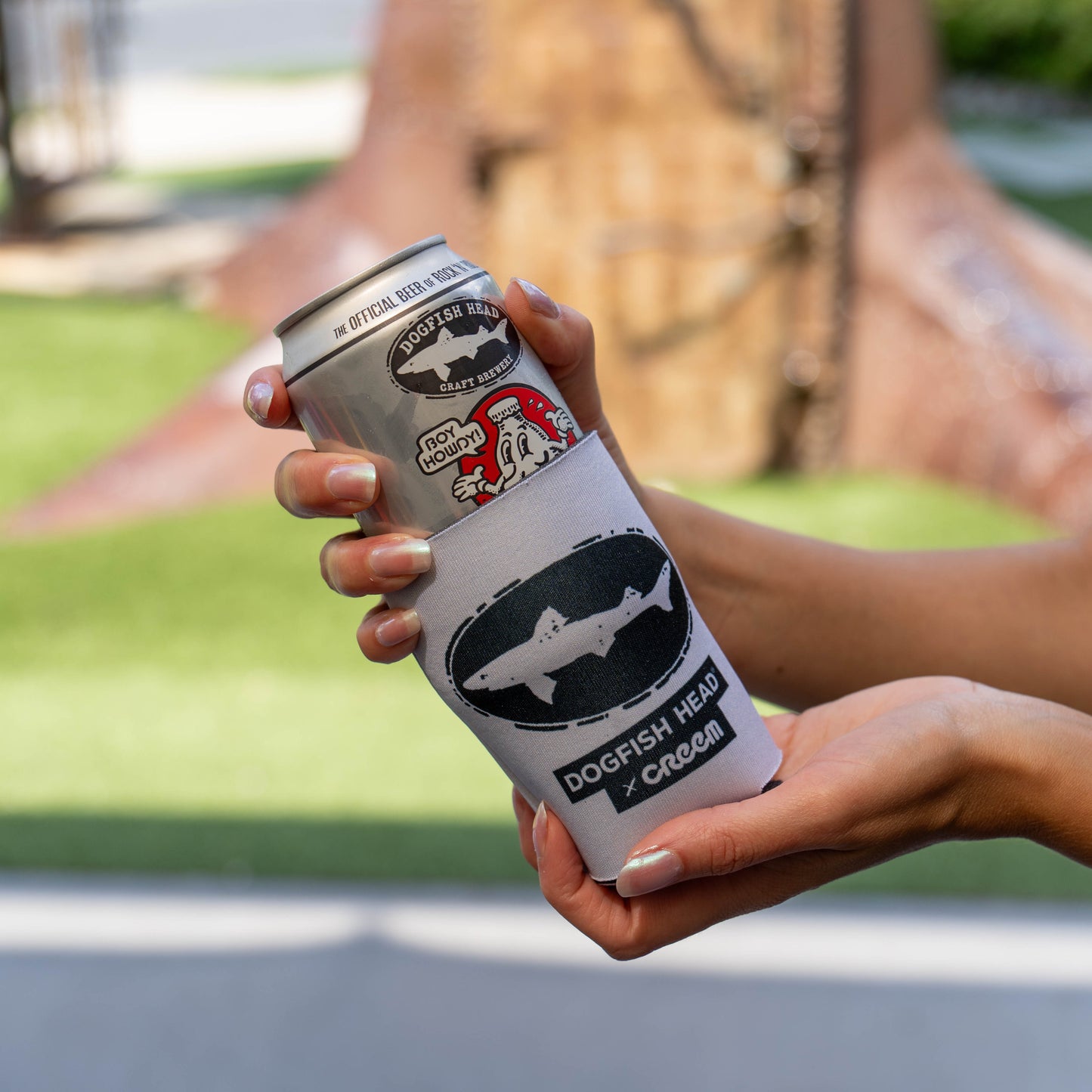 A person holds a silver beverage can with a white dog-eared label featuring a shark logo and the text 'Dogfish Head Cream'. The background shows a blurred outdoor scene with green grass and a brown tree trunk.