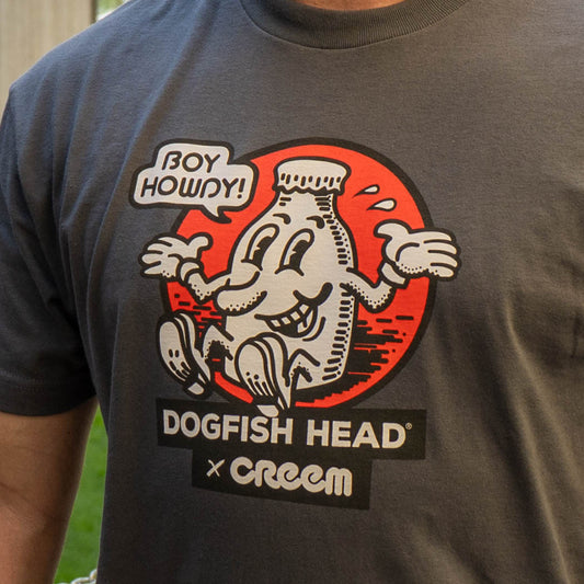 A close-up of a person wearing a dark gray t-shirt featuring a graphic print of the Dogfish Head x Creem collaboration. The graphic shows a white graphic inside a red circle with a speech bubble that reads 'BOY HOWDY!'.