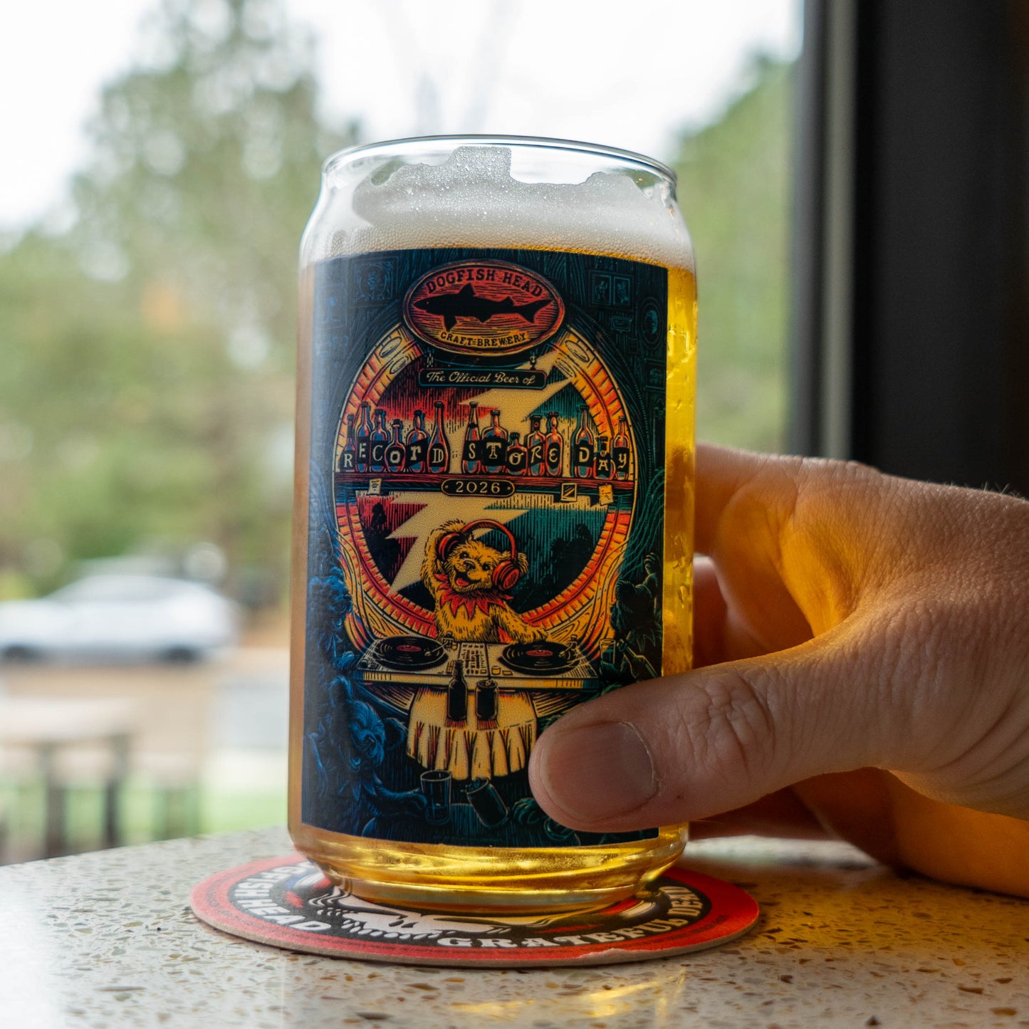 Hand holding Dogfish Head x Grateful Dead 2026 RSD can glass on coaster, highlighting full-color Record Store Day artwork.
