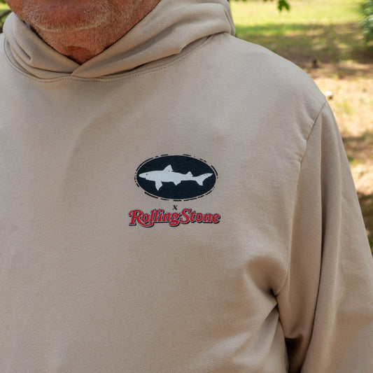 Someone is wearing the Dogfish Head x Rolling Stone Mocha Hoodie by GO WEST T-SHIRT COMPANY, featuring a black oval logo with a white shark and red Rolling Stone text. Grass and trees are visible in the background.