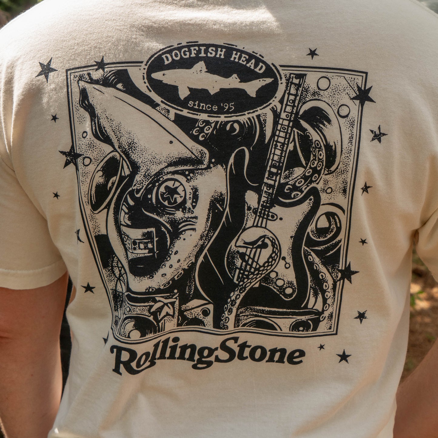 The back of the cream-colored Dogfish Head x Rolling Stone Oatmeal Tee Shirt by GO WEST T-SHIRT COMPANY features a black graphic with a shark, guitar, stars, and "Dogfish Head since 95" and "Rolling Stone.