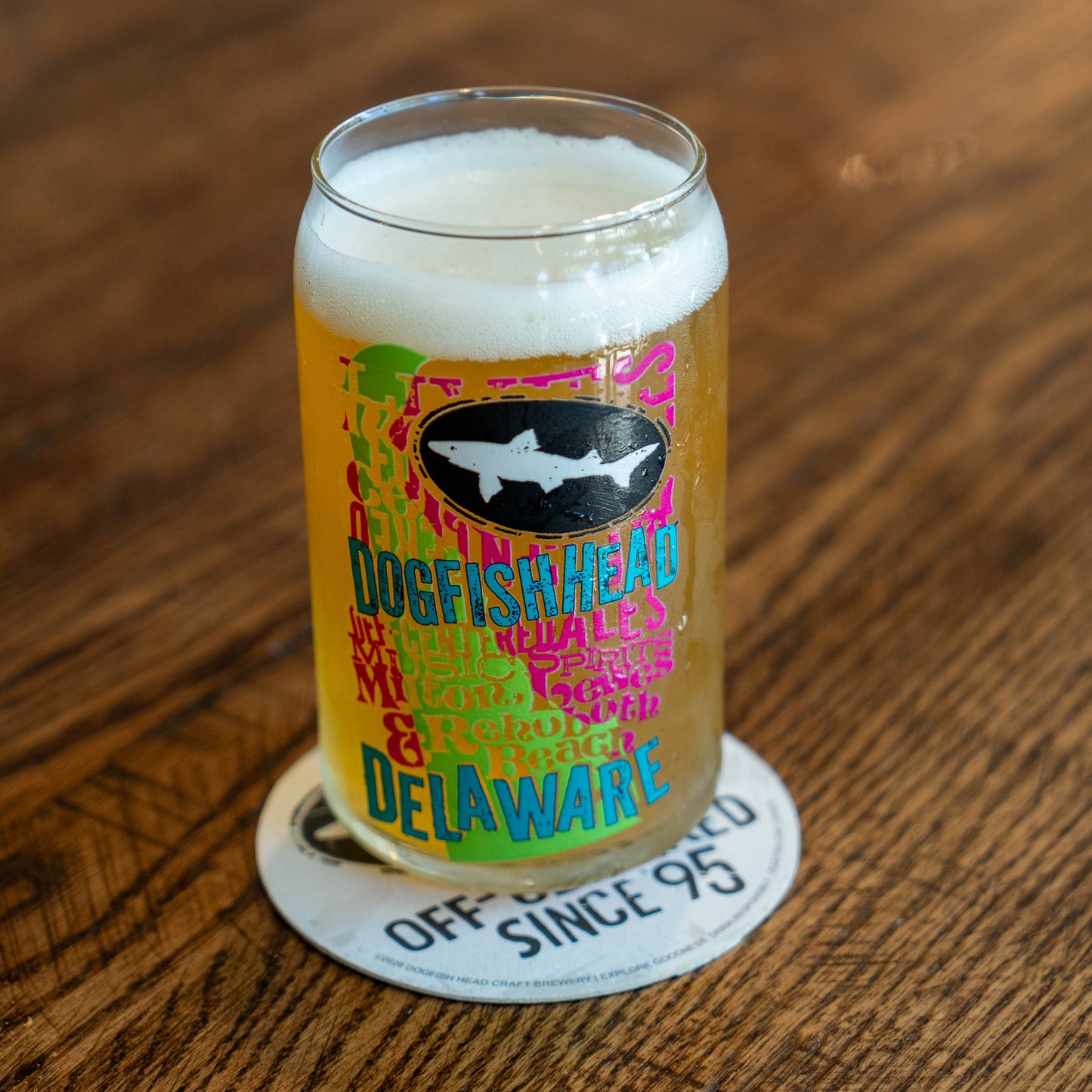 A clear pint glass filled with golden beer and topped with white foam, standing on a round coaster on a wooden table. The glass has a colorful printed design with a shark logo and the words 'Dogfish Head Delaware'.