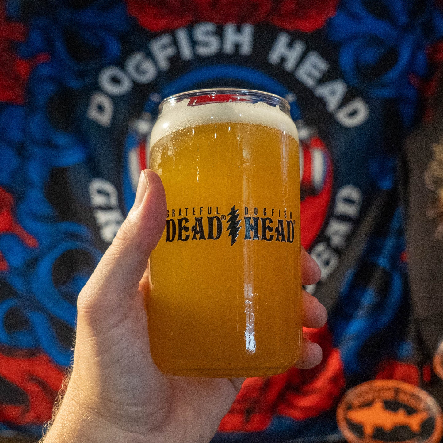 A hand holds a clear glass can-shaped tumbler filled with golden beer, featuring the 'Grateful Dead Dead Head' logo. The background shows a colorful, patterned Dead Head band t-shirt.
