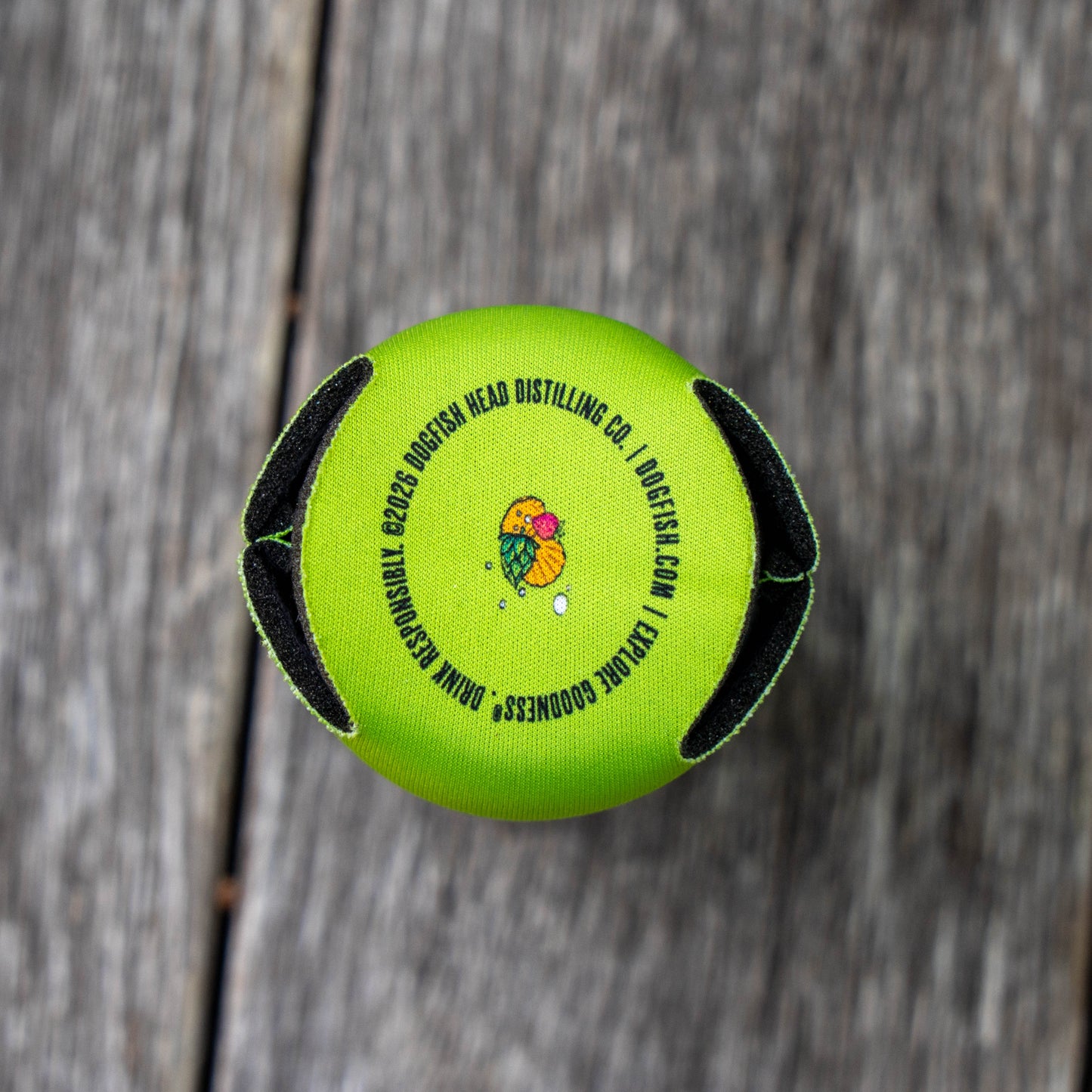Bottom of a bright lime green neoprene can cooler with black foam side panels and a colorful logo in the center, photographed from above on a weathered gray wooden surface.