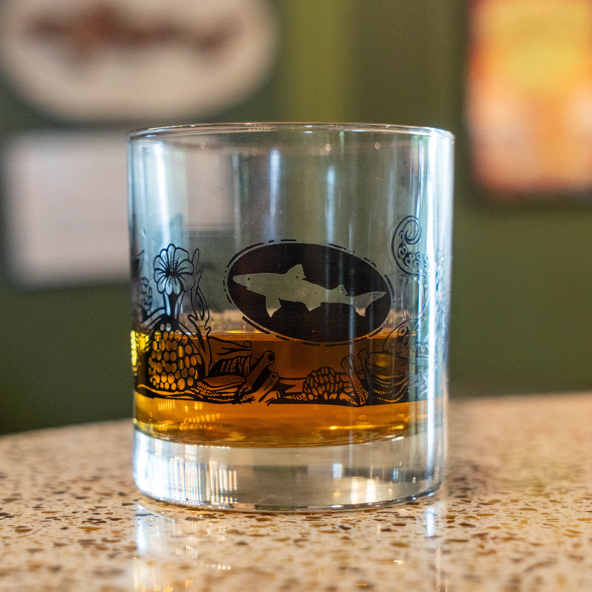The Hull and Helm Rocks Glass by Grandstand Glassware & Apparel rests on a reflective surface, holding amber whiskey. Black octopus tentacle and sea plant illustrations decorate the glass; the background is softly blurred.