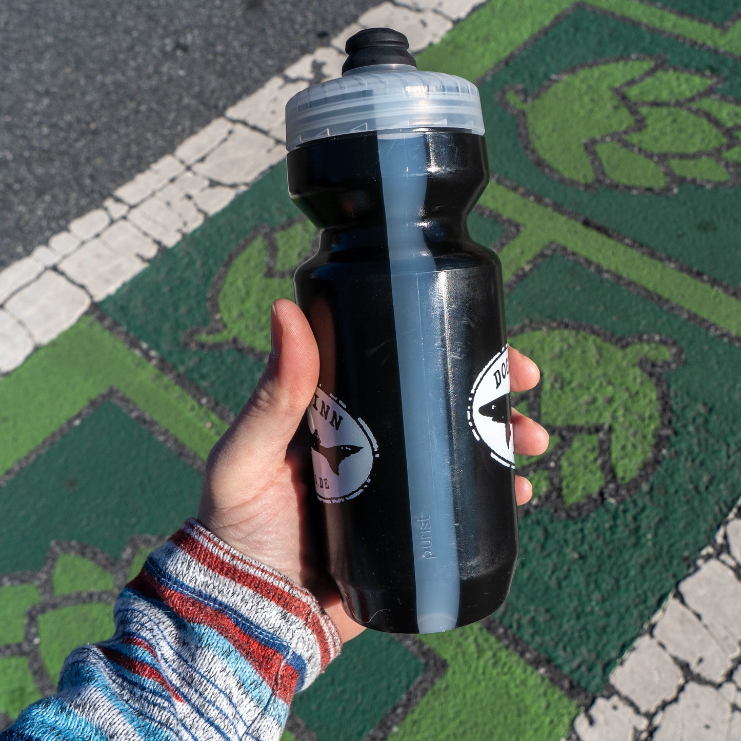 Hand holding a black water bottle with a logo on a road surface