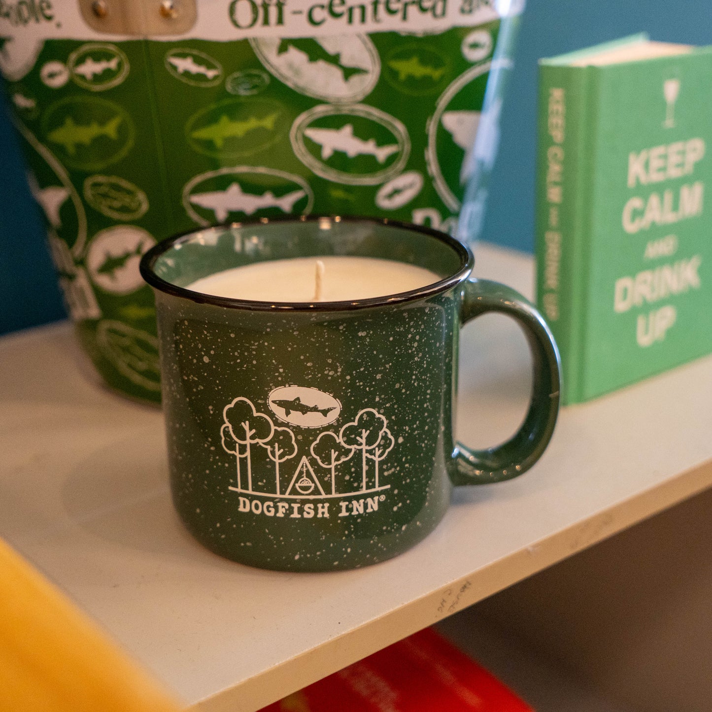 A forest green INN Camp Cup Candle by Dogfish Head Brand sits on a shelf. Behind it, a matching green bucket with white logos and a “KEEP CALM AND DRINK UP” book complete the cozy display.