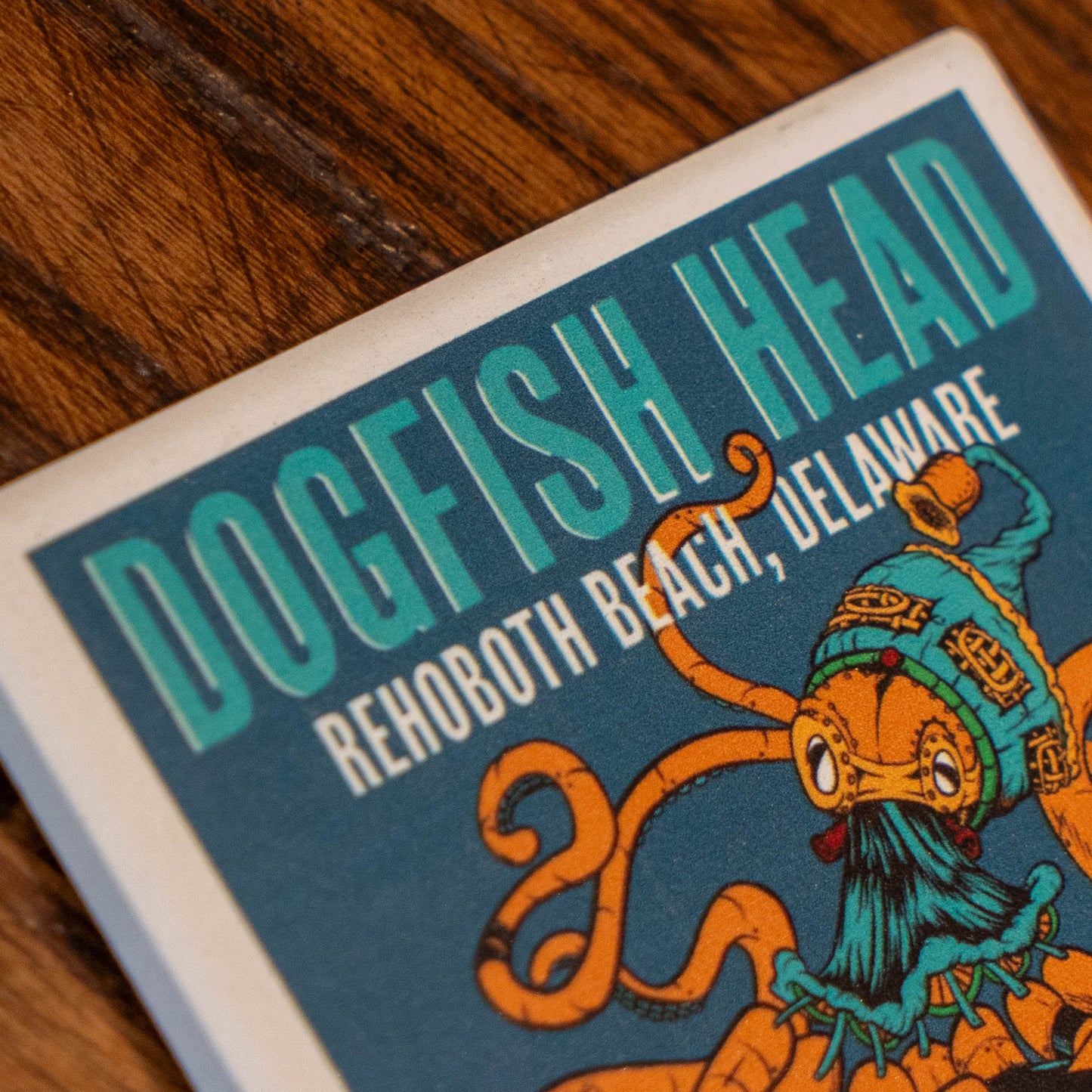 A close-up of a square printed card or coaster featuring the text 'DOGFISH HEAD' and 'REHOBOTH BEACH, DELAWARE' above an illustrated orange octopus wearing a teal knit hat, placed on a wooden surface.