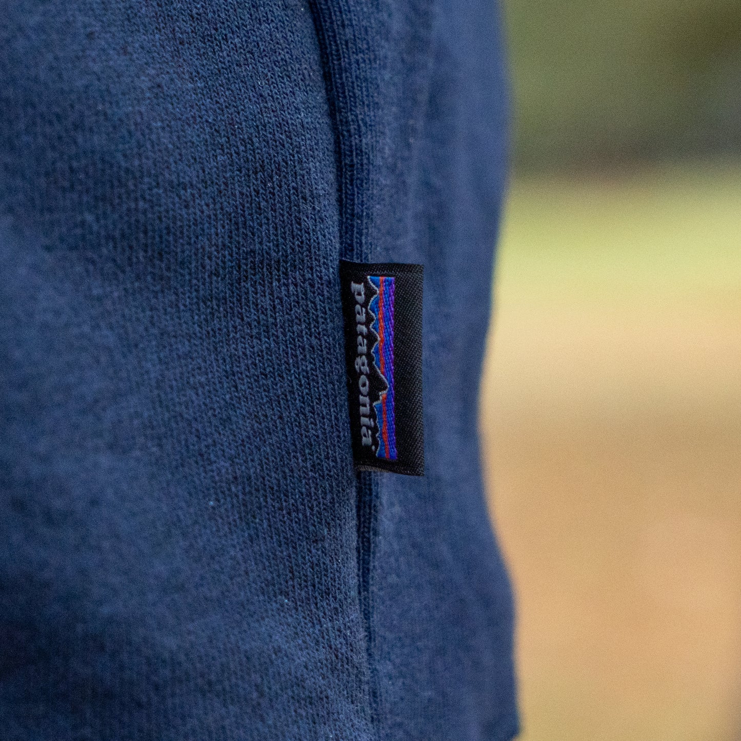 Close-up of the Patagonia Navy Fitz Roy Crew Pullover Sweatshirt with a stitched black tag featuring the colorful mountain logo and white text. The background is softly blurred in neutral shades.