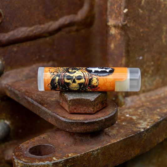 An orange lip balm tube with a black and gold skull graphic sits on a rusty metal bolt against a weathered rusted metal background.