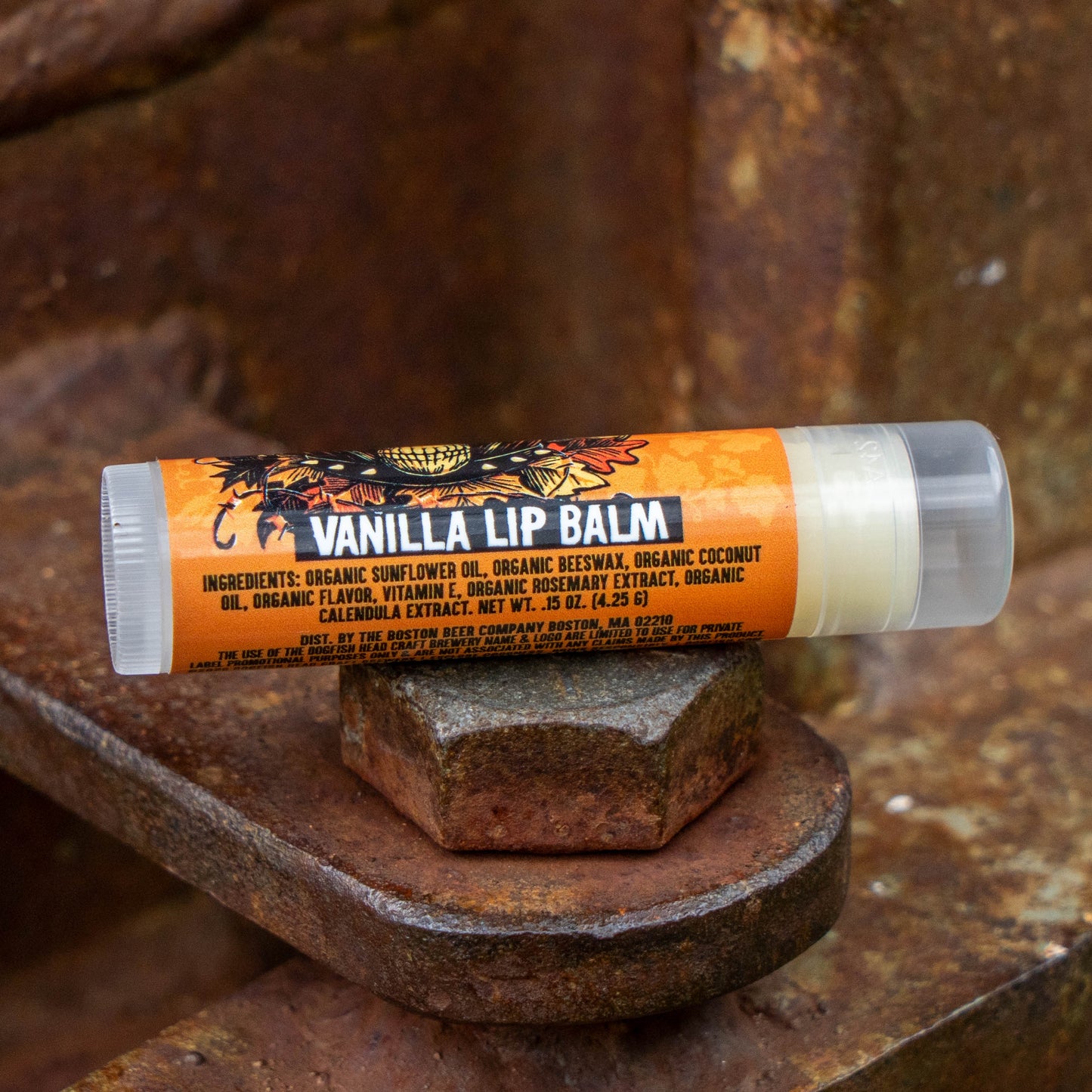 A tube of Vanilla Lip Balm resting on a rustic, weathered metal surface. The lip balm tube features a bright orange label with black text and a floral graphic, and a clear cap showing the white balm inside.
