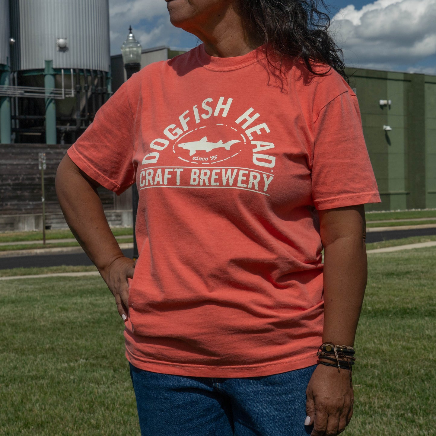 A woman wearing a coral orange t-shirt with a white graphic that reads 'Dogfish Head Craft Brewery' and 'Since 1977' with a shark logo, standing outdoors in front of brewery buildings and a grassy field.