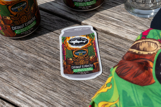 A Dogfish Head x Survivor 50 Bundle sticker by Grandstand Glassware & Apparel on wood, with vibrant fabric and beer cans—perfect for collectors and Survivor fans seeking exclusive merch.