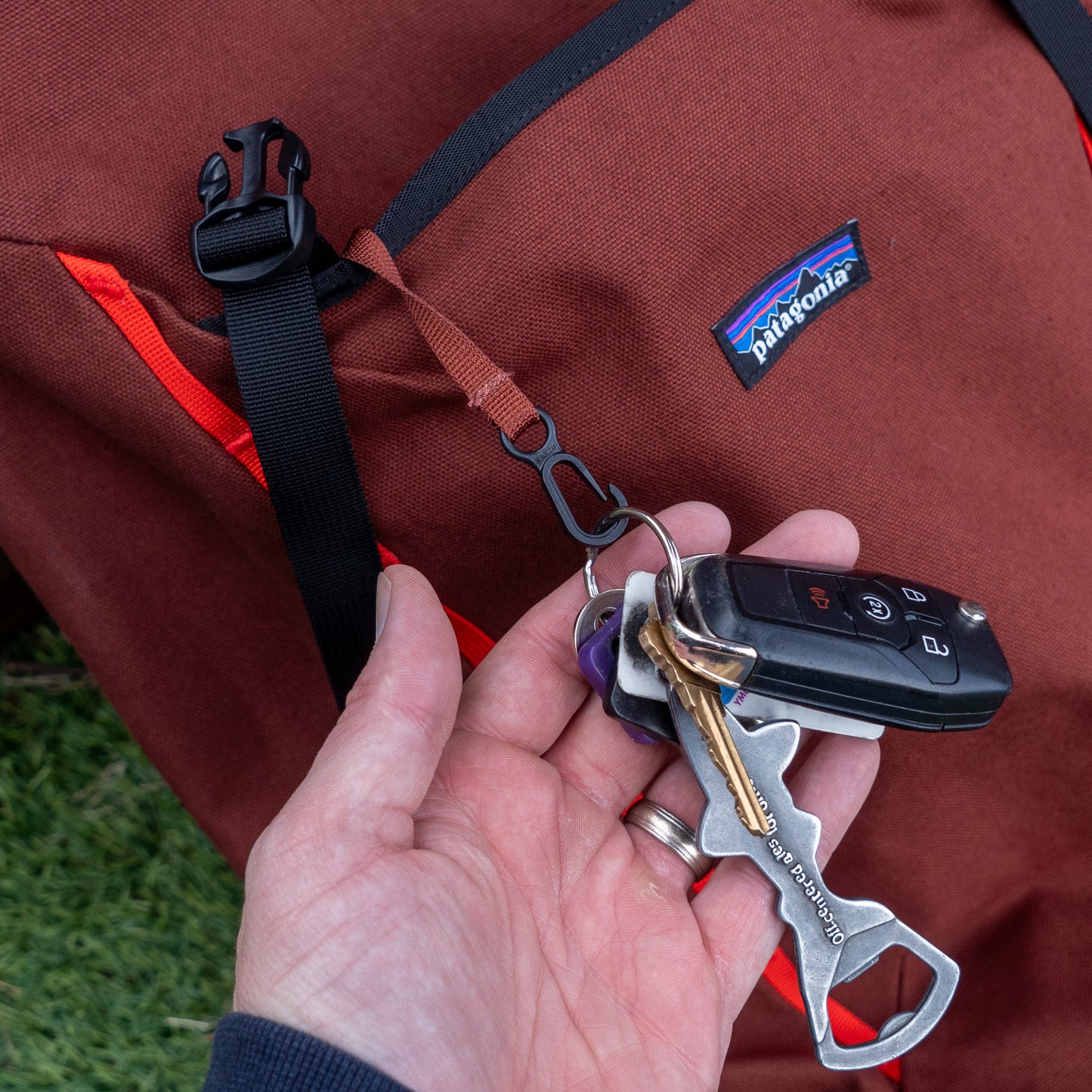 A hand holds keys and a car remote attached to a loop on the brown Patagonia Fieldsmith Lid Pack with a black buckle, resting on grass.