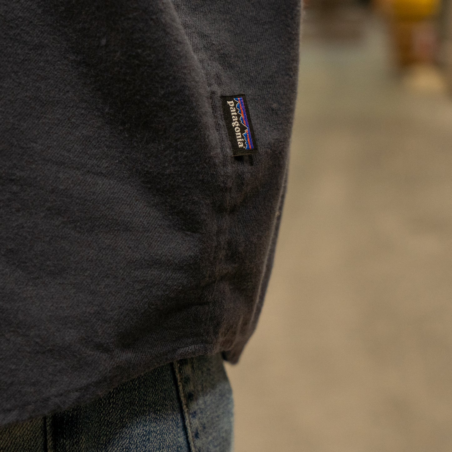 Close-up of a person wearing a Patagonia M Ink Blue Light Weight Flannel Button Up and blue jeans, highlighting the small black Patagonia tag on the shirt’s side seam. The background is blurred.