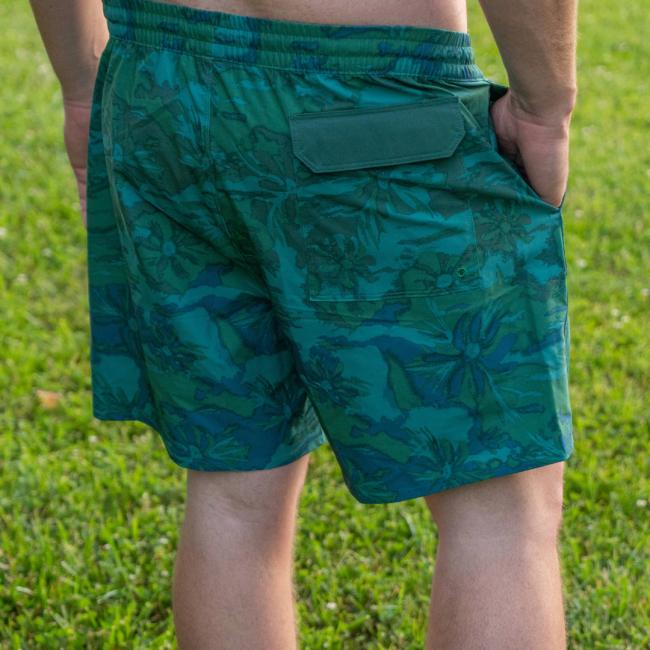 A person standing on grass, waist down, wears Patagonia M Palm Tree Hydropeak Swim Trunks—green and blue floral-print quick-dry swim trunks with a back pocket flap by Patagonia.